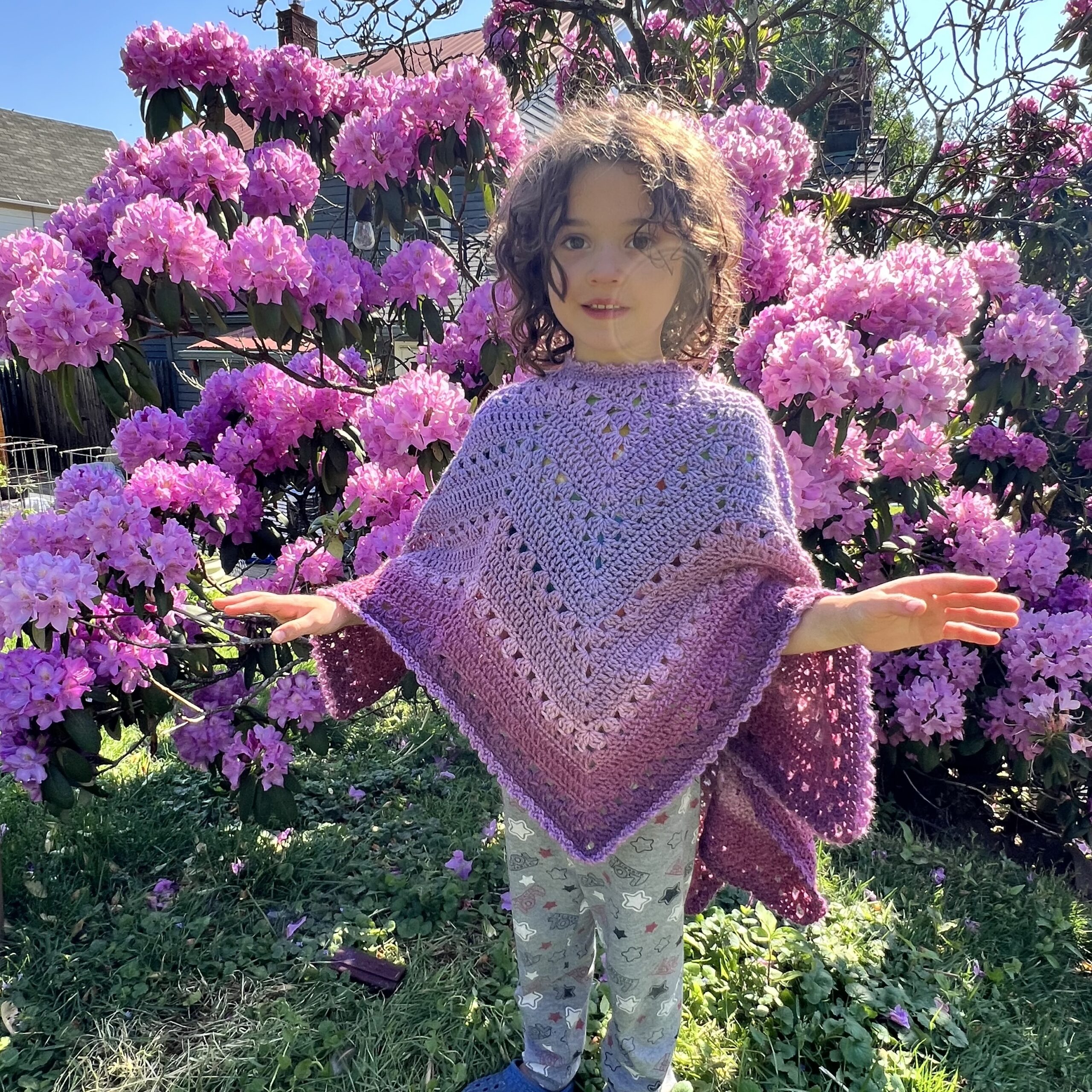 6-Day Star Poncho in Lion Brand Mandala Bonus Bundle Yarn in the color Golum modeled by young child in front of a flowering bush.