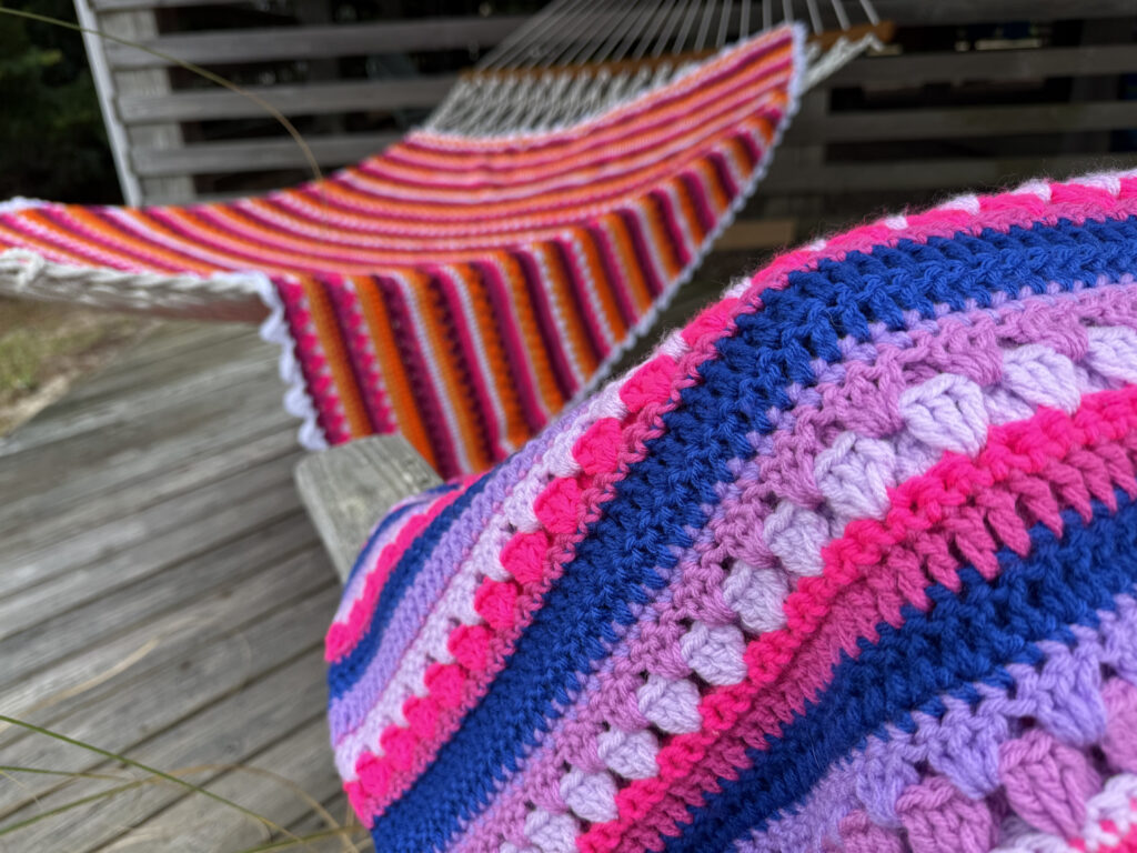This image features two bright, handmade crochet blankets draped over a rustic wooden bench outdoors, set against a lush, green, slightly blurred natural background with trees, grass, and a glimpse of a house or building in the distance. The crochet blankets are the main focal point of the image. It showcases a bold, color palette and intricate stitch work, giving it a rich texture and eye-catching pattern. The crochet pattern is the Retro Rainbow Blanket by Betty McKnit founder of 6-Day Crochet and creator of the viral 6-Day Kid Blanket and 6-Day Star Blanket. The two blankets are made using the colors of the Lesbian pride flag and bi pride flag. The Yarn colors include Hot Pink, Magenta, Lavender, Deep Navy Blue, Orange, Bright Red, and white.