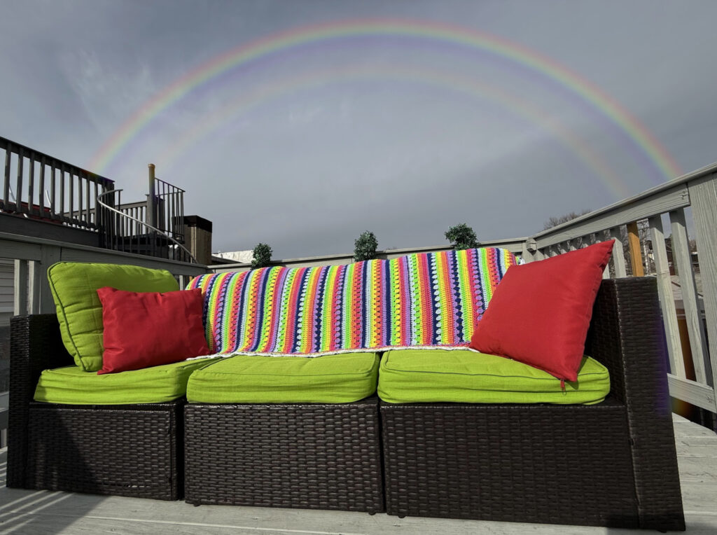 This image shows a colorful crocheted blanket draped over a lime-green cushioned outdoor sofa. The blanket is made up of a repeating stripe pattern using various crochet stitches that create a rich, textured look. The setting appears to be a deck or patio with wooden flooring and a railing in the background. This is the Retro Rainbow Blanket by Betty McKnit founder of 6-Day Crochet and creator of the viral 6-Day Kid Blanket and 6-Day Star Blanket. The yarn colors are extremely vibrant and follow a rainbow-like sequence. Prominent shades include: Pink Purple, in lavender and deeper tones, Blue in Light and Navy, Green in Lime and Grassy hues, A sunny and warm Yellow, Orange, and White used for the scalloped edge. Overall, the image conveys a cozy, inviting, and cheerful atmosphere—ideal for relaxing outdoors with a handmade touch of warmth and color.