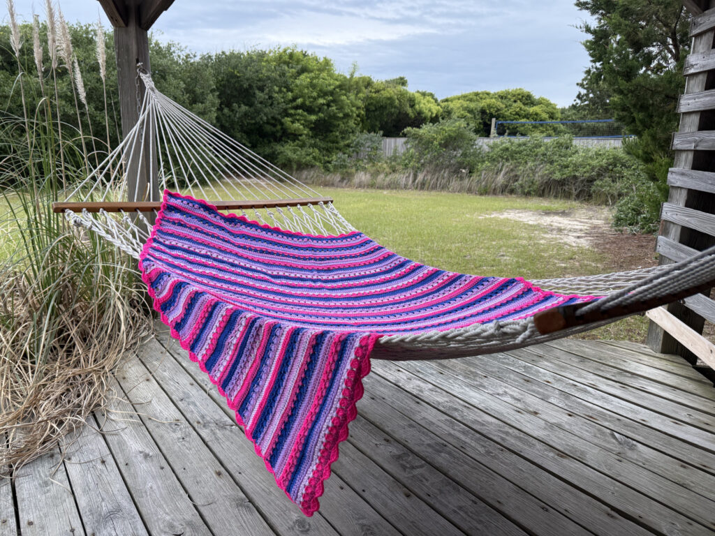 This image captures a vibrant handmade crochet blanket draped over a white rope hammock in a serene, outdoor setting. The crochet blanket is crafted with alternating rows of vivid colors including hot pink, lavender, navy blue, and light purple. The edge is finished with a scalloped border in bright pink, adding a soft and decorative touch that enhances its overall aesthetic appeal. The Crochet Pattern for this blanket is the Retro Rainbow Crochet Pattern by Betty McKnit founder of 6-Day Crochet and creator of the viral 6-Day Kid Blanket and 6-Day Star Blanket. Each row is carefully color-coordinated, creating a Bi Pride Flag-inspired motif. The blanket is spread in a relaxed fashion, suggesting comfort and leisure, evoking a cozy, tranquil vibe perfect for lounging outdoors.