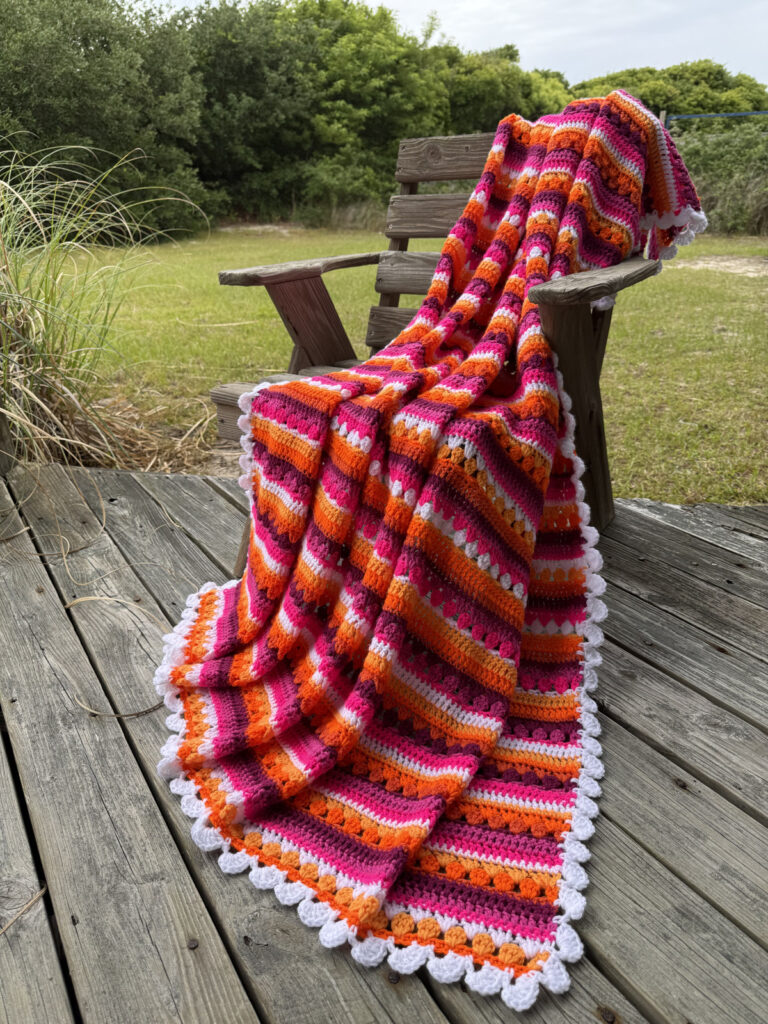 This image features a handmade crochet blanket draped gracefully over a rustic wooden bench in an outdoor setting. The blanket’s rich colors and detailed stitch work are captured beautifully, offering a vibrant contrast against the muted, natural tones of the background. The Crochet Pattern for this blanket is the Retro Rainbow Crochet Pattern by Betty McKnit founder of 6-Day Crochet and creator of the viral 6-Day Kid Blanket and 6-Day Star Blanket. Each row is carefully color-coordinated, creating a Lesbian Pride Flag-inspired motif. The blanket is finished in a scalloped edge trim, which adds a decorative, soft finish in white. The color palette includes bold and feminine tones such as hot pink, orange, dark magenta, and white accent rows. The color pattern is symmetrical, giving a sense of order and intentional design.