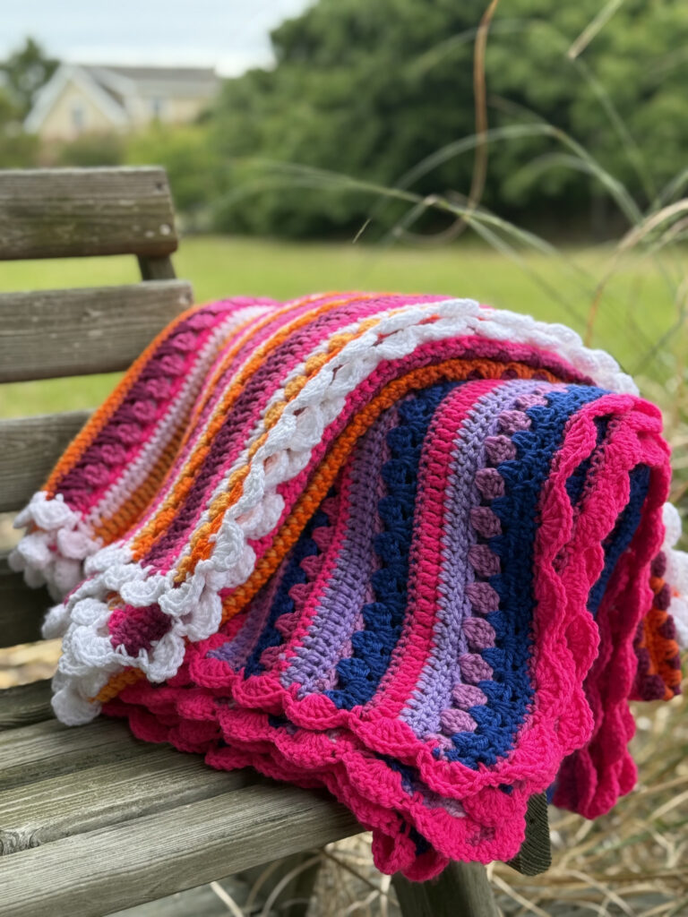 This image features two bright, handmade crochet blankets draped over a rustic wooden bench outdoors, set against a lush, green, slightly blurred natural background with trees, grass, and a glimpse of a house or building in the distance. The crochet blankets are the main focal point of the image. It showcases a bold, color palette and intricate stitch work, giving it a rich texture and eye-catching pattern. The crochet pattern is the Retro Rainbow Blanket by Betty McKnit founder of 6-Day Crochet and creator of the viral 6-Day Kid Blanket and 6-Day Star Blanket. The two blankets are made using the colors of the Lesbian pride flag and bi pride flag. The Yarn colors include Hot Pink, Magenta, Lavender, Deep Navy Blue, Orange, Bright Red, and white.