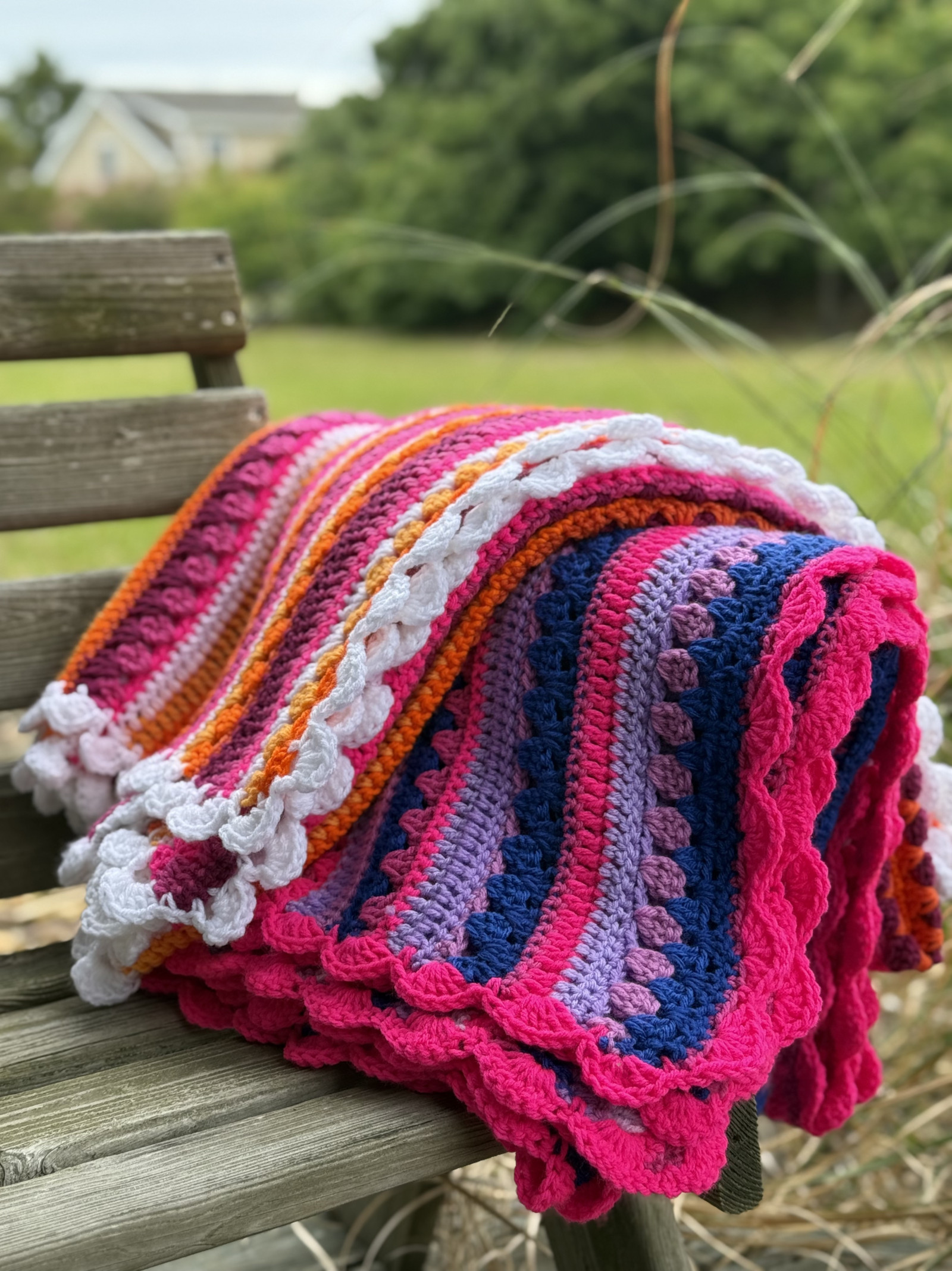 This image features two bright, handmade crochet blankets draped over a rustic wooden bench outdoors, set against a lush, green, slightly blurred natural background with trees, grass, and a glimpse of a house or building in the distance. The crochet blankets are the main focal point of the image. It showcases a bold, color palette and intricate stitch work, giving it a rich texture and eye-catching pattern. The crochet pattern is the Retro Rainbow Blanket by Betty McKnit founder of 6-Day Crochet and creator of the viral 6-Day Kid Blanket and 6-Day Star Blanket. The two blankets are made using the colors of the Lesbian pride flag and bi pride flag. The Yarn colors include Hot Pink, Magenta, Lavender, Deep Navy Blue, Orange, Bright Red, and white.