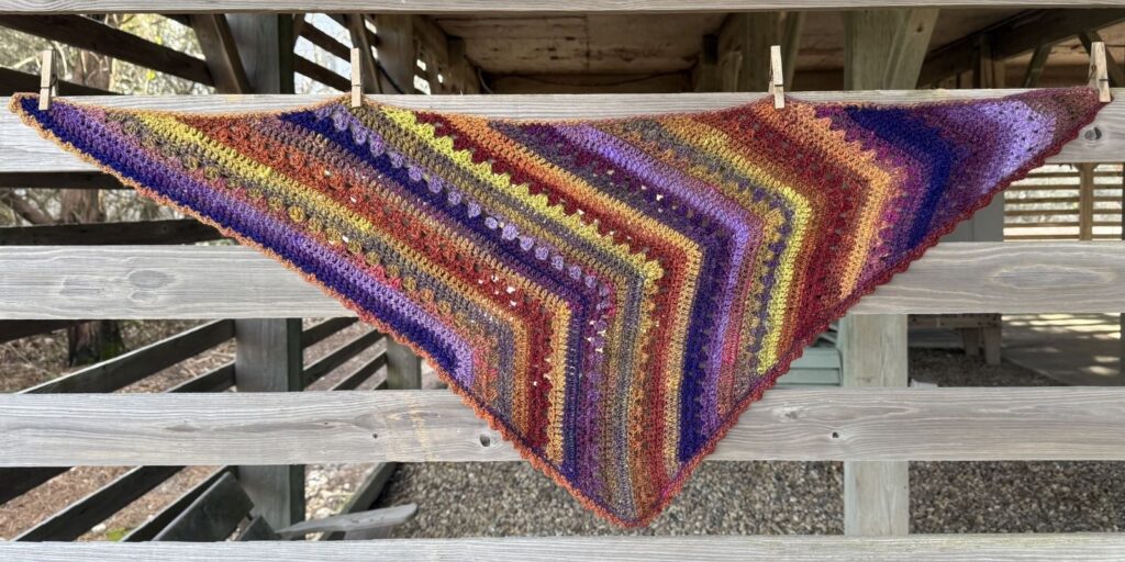 Colorful 6-Day crochet scarf hanging on wooden siding.