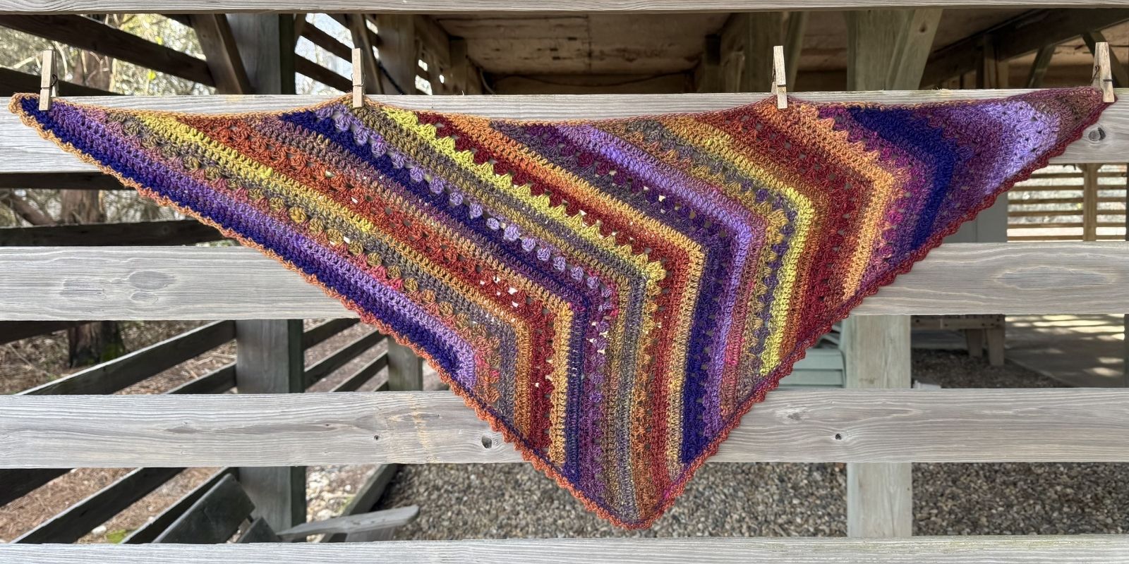 Colorful 6-Day crochet scarf hanging on wooden siding.