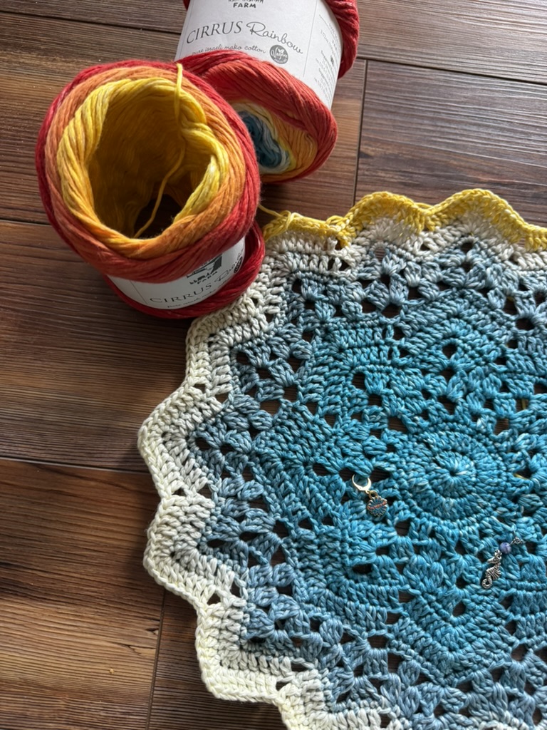 A circular crochet start of a star blanket in shades of deep teal, light blue, and creamy white rests on a wooden floor alongside two yarn cakes with a vibrant red, orange, and yellow color gradient, with small stitch markers attached to the doily.