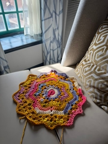 A circular, mandalalike crochet piece in progress with a colorful palette transitioning from a bright pink and white center through shades of yellow, orange, pink, purple, and blue.