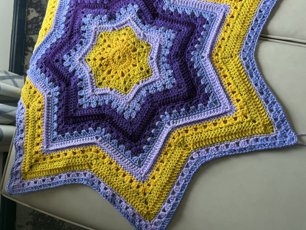 A vibrant, star-shaped crochet blanket featuring concentric rings of yellow, light purple, dark purple, and periwinkle blue, resting on a light-colored upholstered surface.