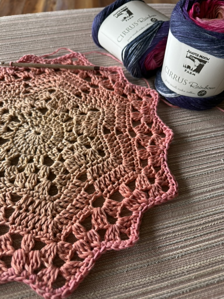 A circular crochet blanket in progress with a gradient from creamy white in the center to warm orange, pink, and brown, featuring an adjacent ball of "Cirrus Rainbow" yarn and a crochet hook resting on a striped upholstered surface.