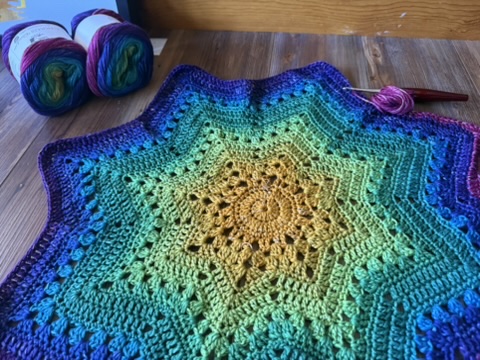 A large, star-shaped crochet project featuring vibrant rainbow stripes transitioning from yellow and green through deep teal to blue and purple, set next to two yarn cakes on a wooden floor.